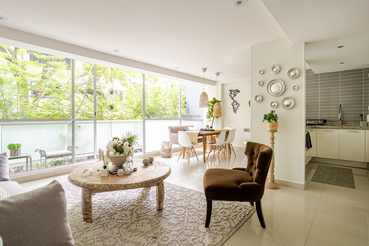 An open living space features large glass windows allowing natural light to fill the room. A round wooden coffee table is central, accompanied by a comfortable armchair. A dining table for four is visible, along with a modern kitchen area in the background.