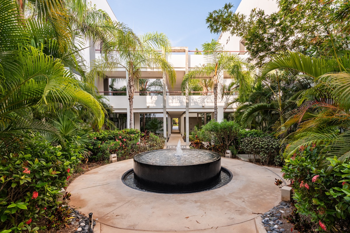 A tranquil courtyard is centered around a round fountain, surrounded by lush greenery and vibrant flowers. The pathway leads through the manicured landscaping, showcasing the peaceful atmosphere of the residential area, highlighted by the soft sounds of flowing water.