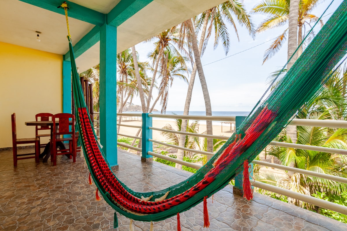 A spacious balcony overlooks the beach, featuring a colorful hammock and a wooden dining set with four chairs. Palm trees frame the view, and the ocean is visible in the distance, creating an inviting outdoor space for relaxation.