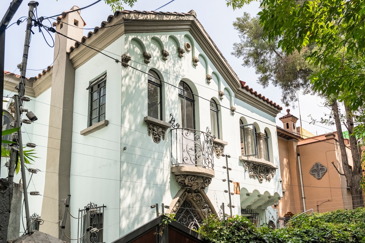 The exterior of a unique two-story house features pastel blue walls and decorative architectural details, including arched windows and a wrought iron balcony. Lush greenery surrounds the property, blending with the vibrant atmosphere of the neighborhood.