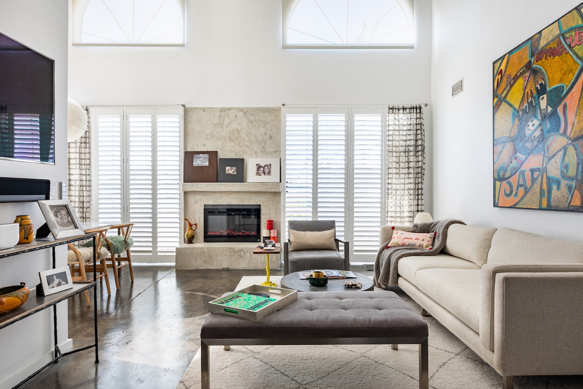 A bright living area features high ceilings and large windows with modern shutters. A comfortable cream sofa and a plush gray ottoman center the space, accompanied by a minimalist coffee table. An artistic wall accent adds color, while a linear fireplace enhances the ambiance.