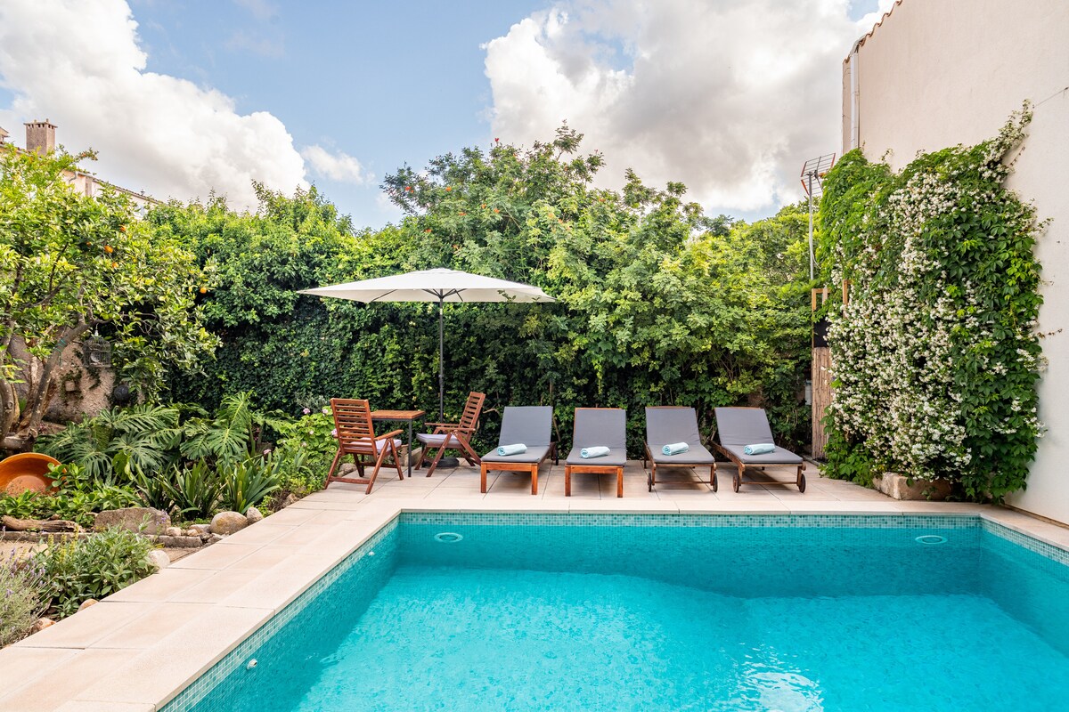 A serene saltwater pool is surrounded by lush greenery and tall trees, creating a private oasis. Six comfortable sun loungers are arranged beside the pool, accompanied by a patio table and chairs shaded by a large umbrella, inviting relaxation.