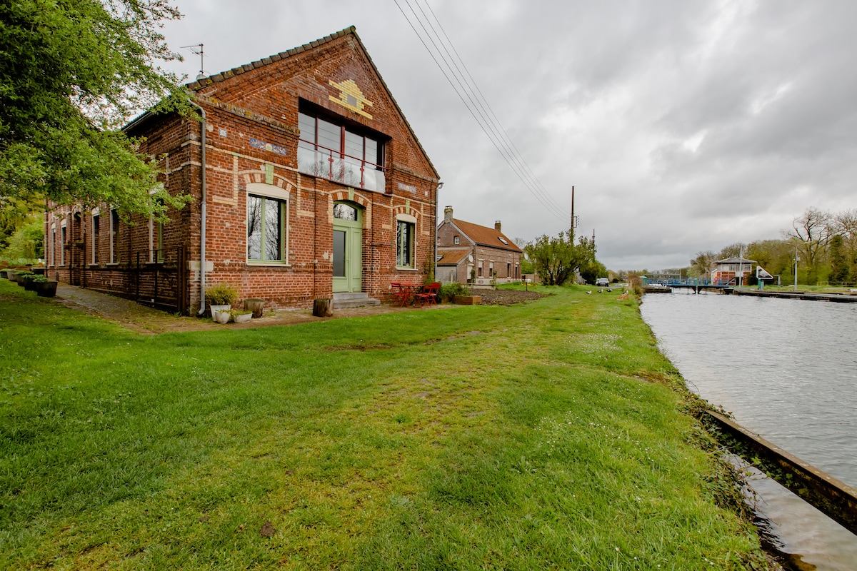A charming brick building is situated near a peaceful canal, surrounded by well-maintained green grass. Two large windows with white curtains frame the entrance, while red seating is visible on the patio. The serene waters of the canal reflect the overcast sky.