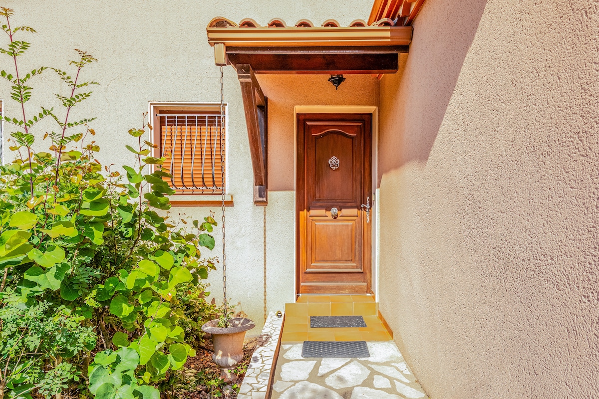 An entrance to the villa is framed by lush greenery and a well-maintained garden. The wooden door, adorned with a decorative knocker, invites guests inside. A welcoming stone pathway leads to the door, complemented by a small, decorative pot placed nearby.