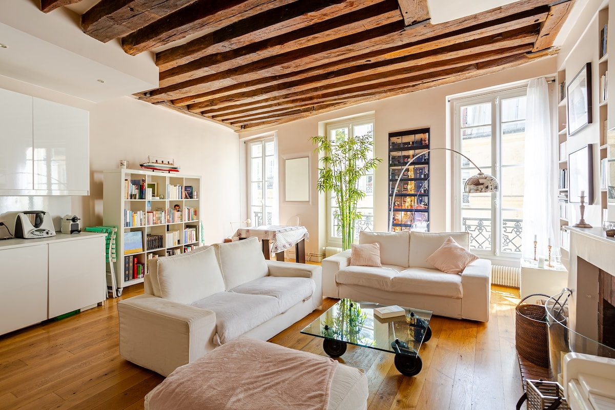 The living room features exposed wooden beams and large windows providing ample natural light. Two sofas face each other, accompanied by a glass coffee table. A bookshelf filled with books and a potted plant enhance the space, while a modern arc lamp adds a stylish touch.