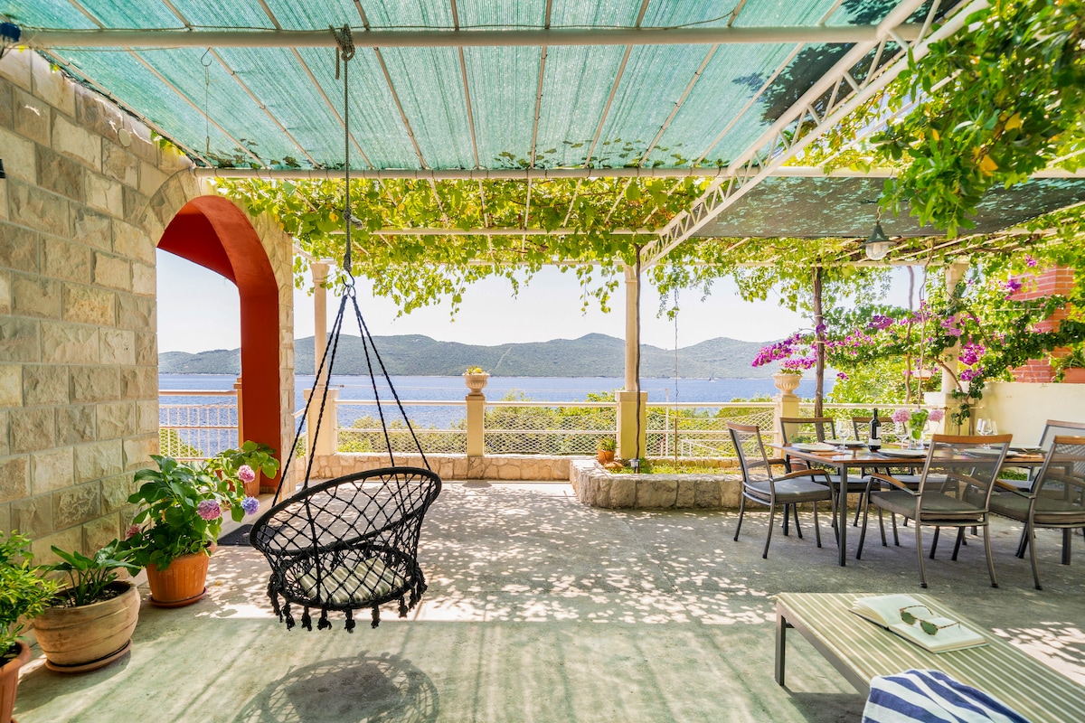 An outdoor terrace features a hanging swing chair surrounded by potted plants. A dining table is set under a pergola draped with vines, offering views of the sea and distant hills. Sunlight filters through the canopy, creating a serene atmosphere.