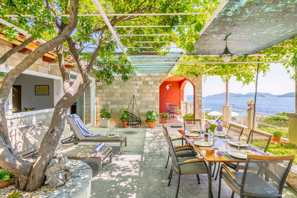 A shaded outdoor terrace features a dining area with chairs and a large table, surrounded by potted plants. Sun loungers are positioned nearby, with a view of the sea visible through the decorative pergola overhead. The tranquil atmosphere is enhanced by the greenery.