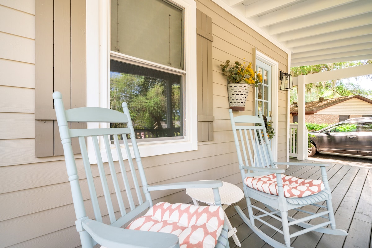 A welcoming front porch features two light blue rocking chairs with coral-patterned cushions. A small white table rests between them, while a window adorned with shutters provides a view into the cottage. Bright yellow flowers are arranged in a hanging basket nearby.