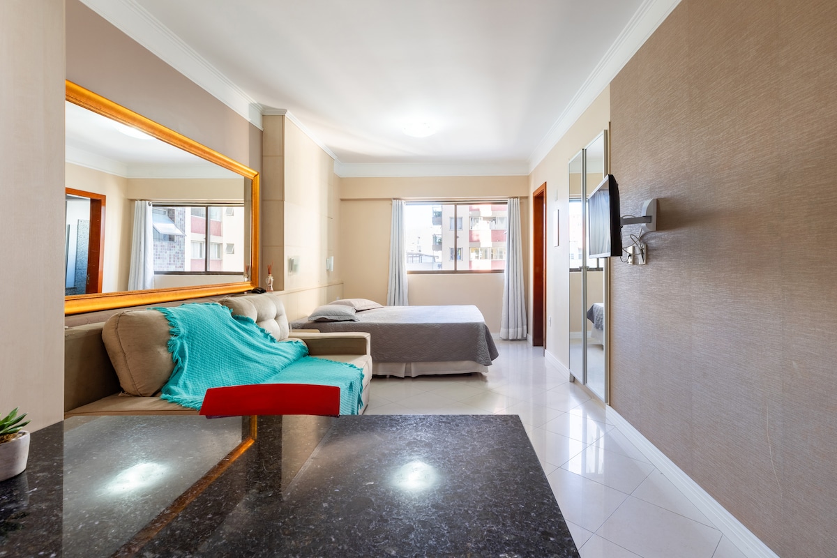 A combined living space and bedroom area is visible, featuring a spacious bed and a sofa. A large mirror and a flat-screen TV are mounted on the wall, while natural light enters through the window. A dining area is positioned in the foreground with a dark countertop.