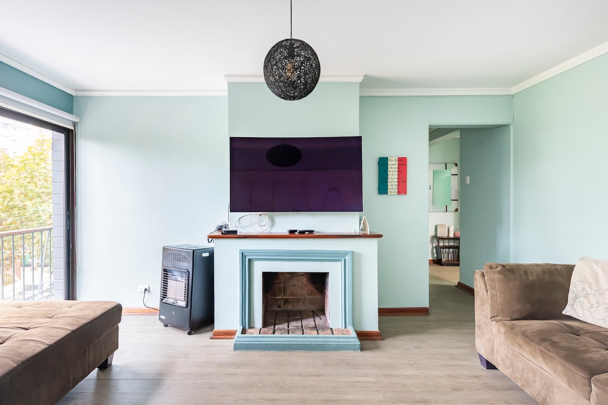 A living area is showcased with a light blue wall and wooden flooring. A modern fireplace is positioned beneath a wall-mounted television. Two brown sofas are arranged for seating, with a decorative pendant light hanging overhead, providing a cozy atmosphere for relaxation.