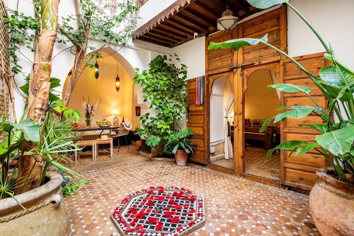 A central courtyard features a traditional mosaic pattern on the floor, surrounded by lush green plants and artisanal pottery. Wooden doorways lead to adjacent rooms, while decorative lanterns hang from the ceiling, adding a touch of warmth to the inviting space.