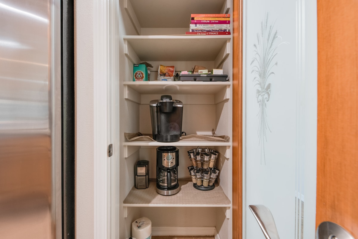 A well-organized pantry is displayed with multiple shelves. On the lower level, a coffee maker and a single-serve machine are visible, alongside a variety of coffee pods. Cookbooks and assorted containers are arranged above, providing easy access to kitchen essentials.