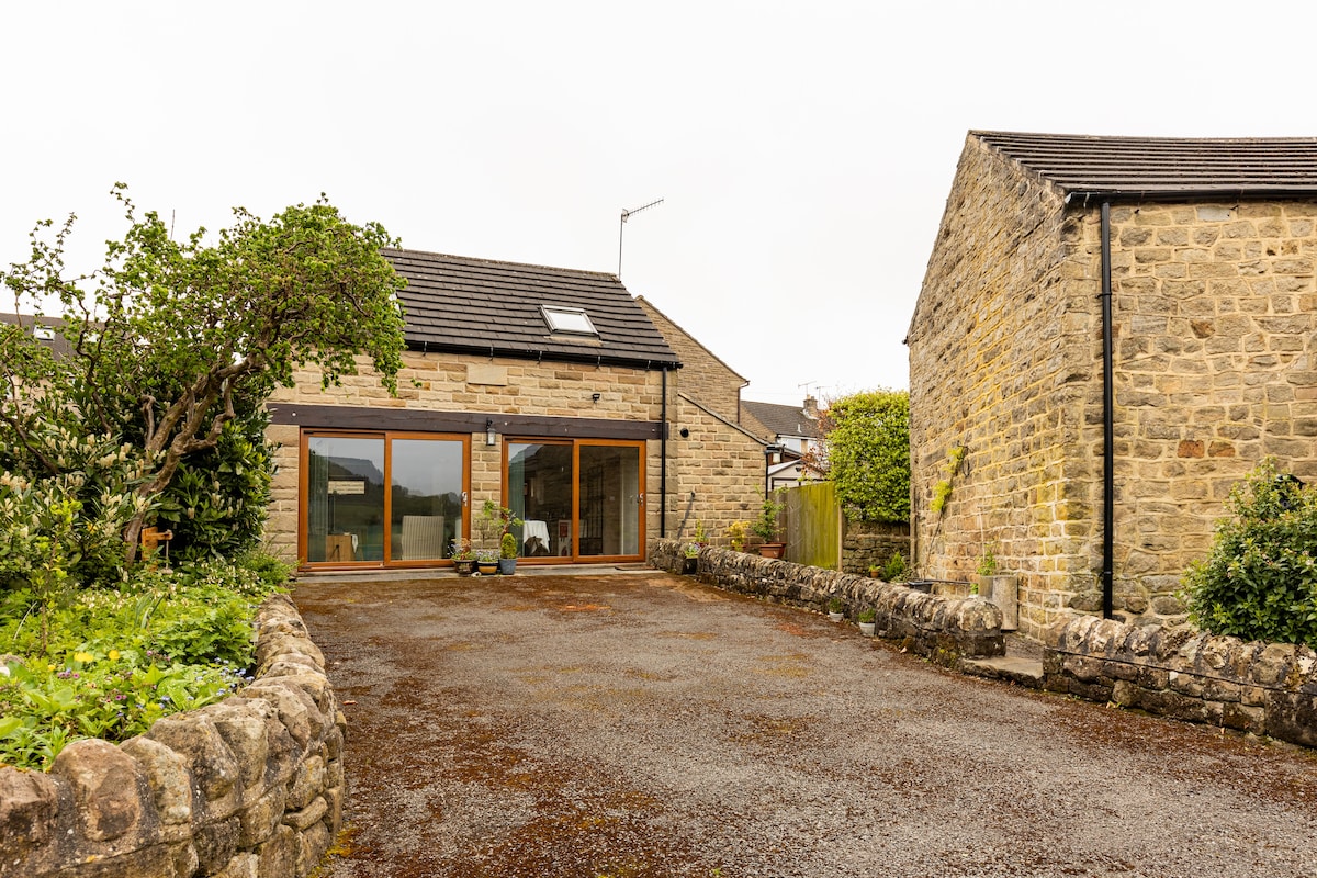 A modern holiday home is depicted with large glass doors offering a view of the surrounding area. The exterior features a blend of natural stone and contemporary elements. A spacious gravel area is visible in front, bordered by low stone walls and greenery.