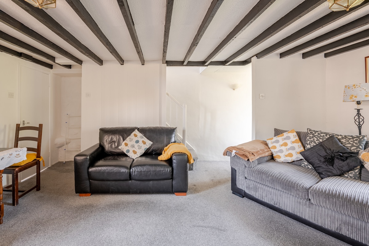 The lounge area is furnished with two comfortable sofas, one in black leather and the other in a vertical striped pattern, adorned with cushions. A small dining table is positioned nearby, and soft lighting is provided by two ceiling fixtures that highlight the original beams above.