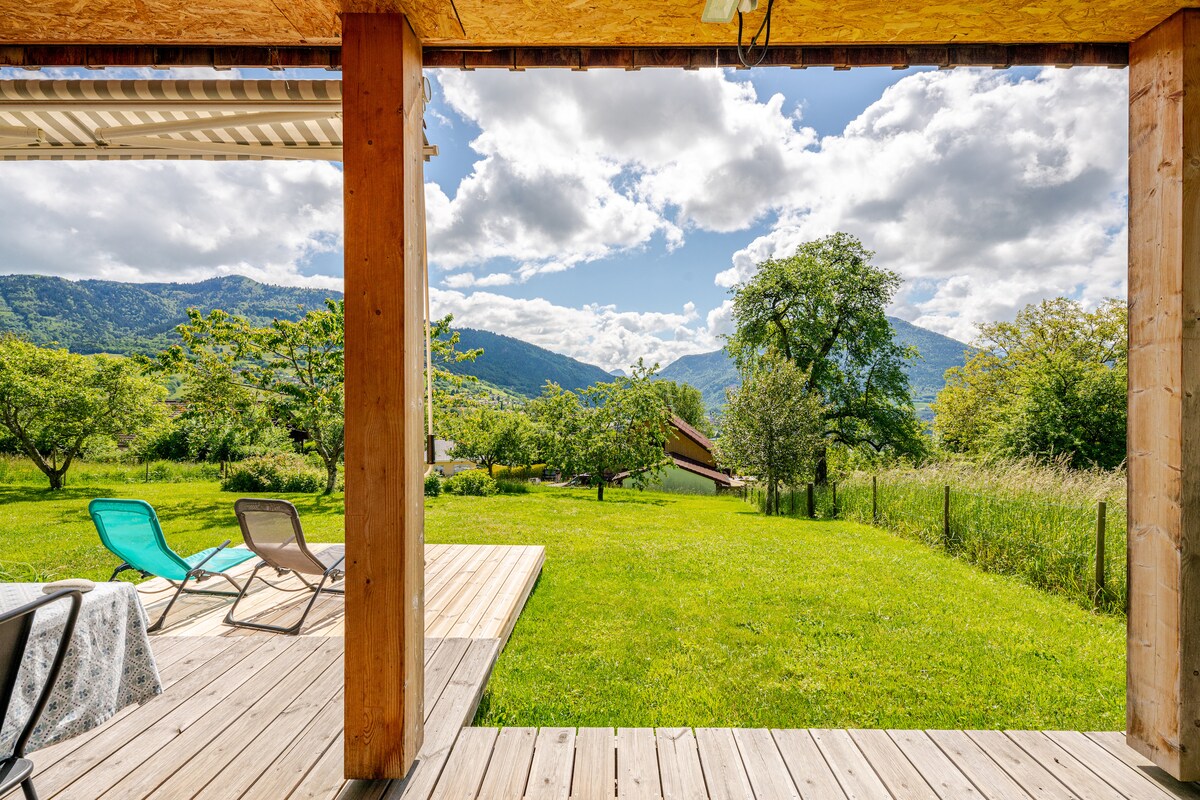 A spacious wooden terrace is visible, framed by sturdy support beams. Green grass stretches out towards distant mountains, under a partly cloudy sky. Two lounge chairs invite relaxation, while trees provide a natural backdrop, enhancing the serene outdoor space.