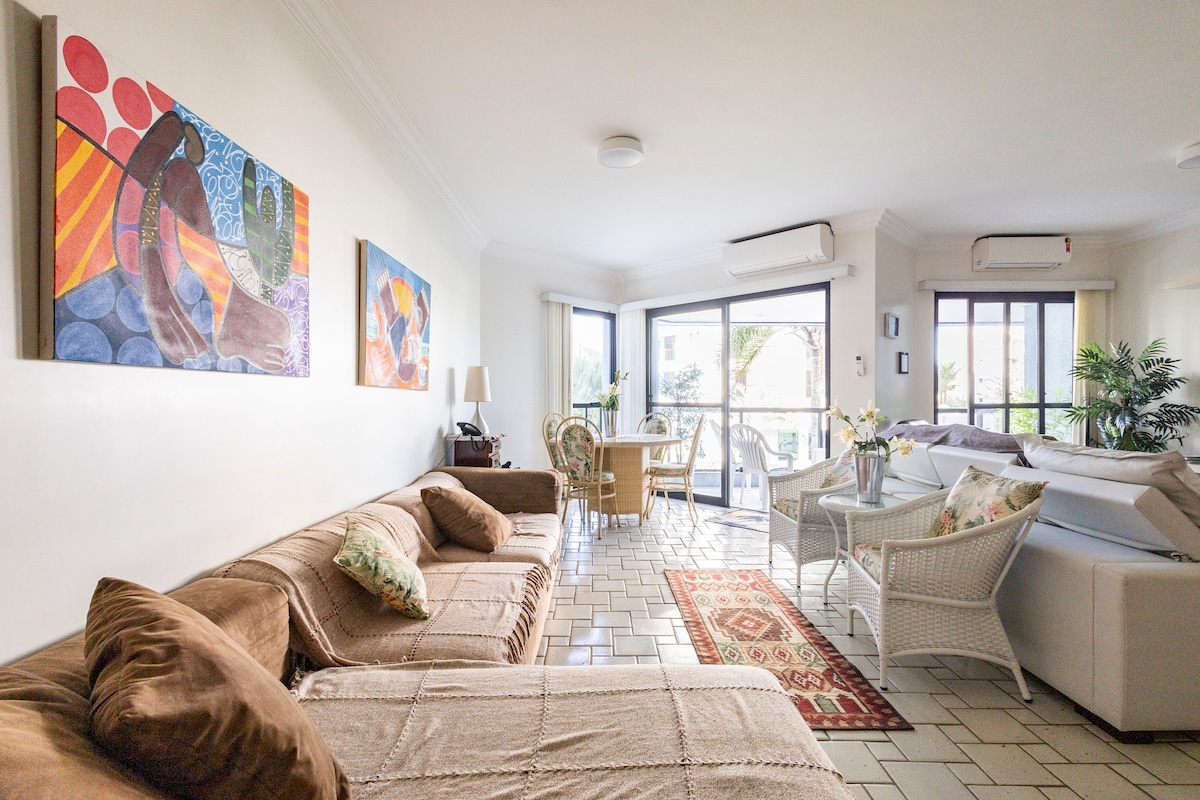 A spacious living area is showcased with large windows allowing natural light. Soft furnishings, including a sectional sofa and wicker chairs, are arranged around a decorative rug. Colorful artwork adorns the walls, creating a friendly and inviting space for relaxation.