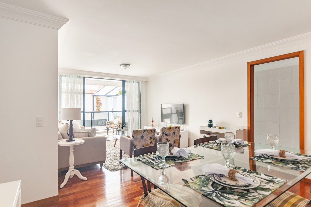 A dining area is presented with an elegant glass table set for four. The space is illuminated by natural light from large windows that open to a comfortable living area featuring a plush sofa and a TV. Colorful accents add a touch of style.