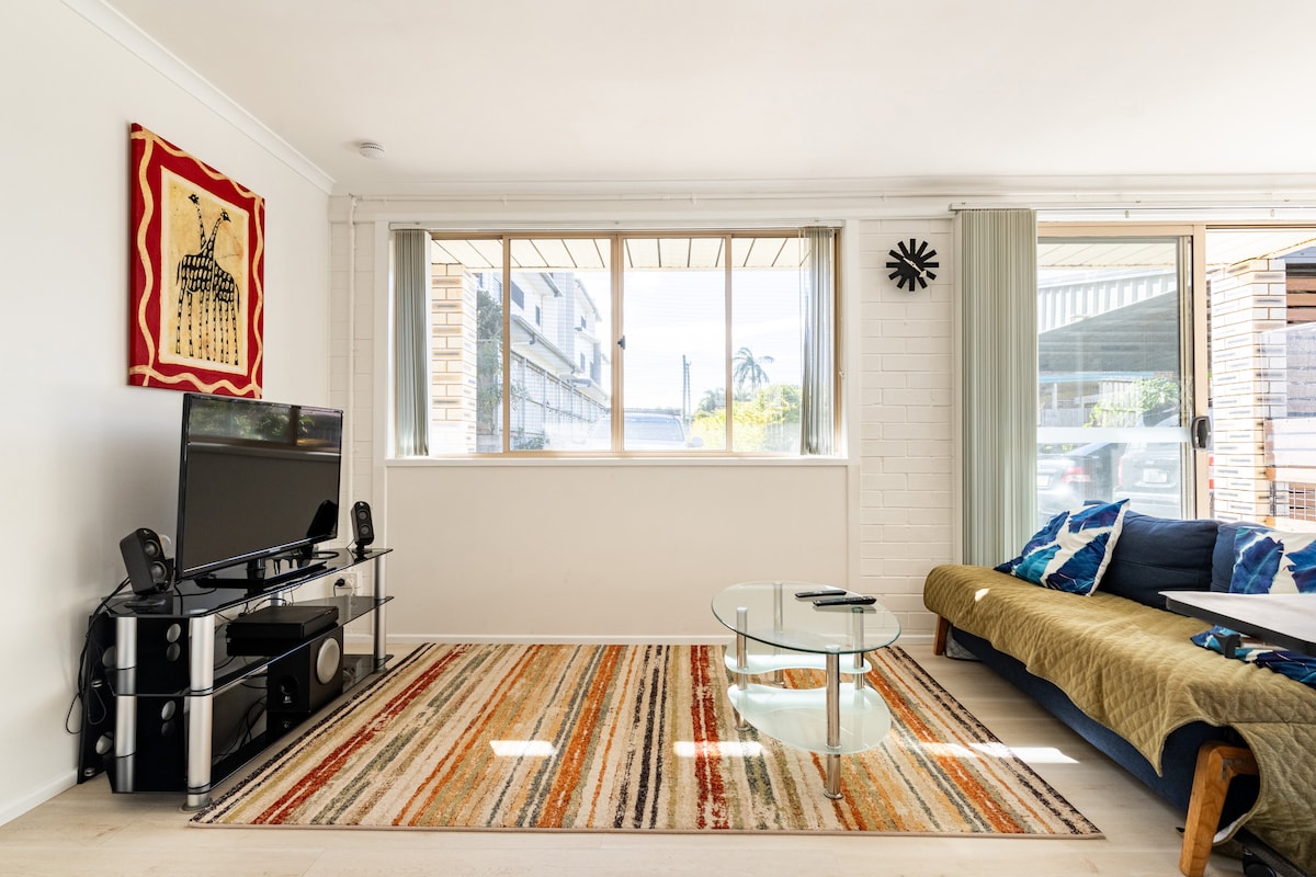 A sitting area is presented with a glass coffee table positioned on a colorful striped rug. A sofa in muted tones is paired with a vibrant wall hanging. Large windows allow natural light to fill the space, creating a bright and airy atmosphere.