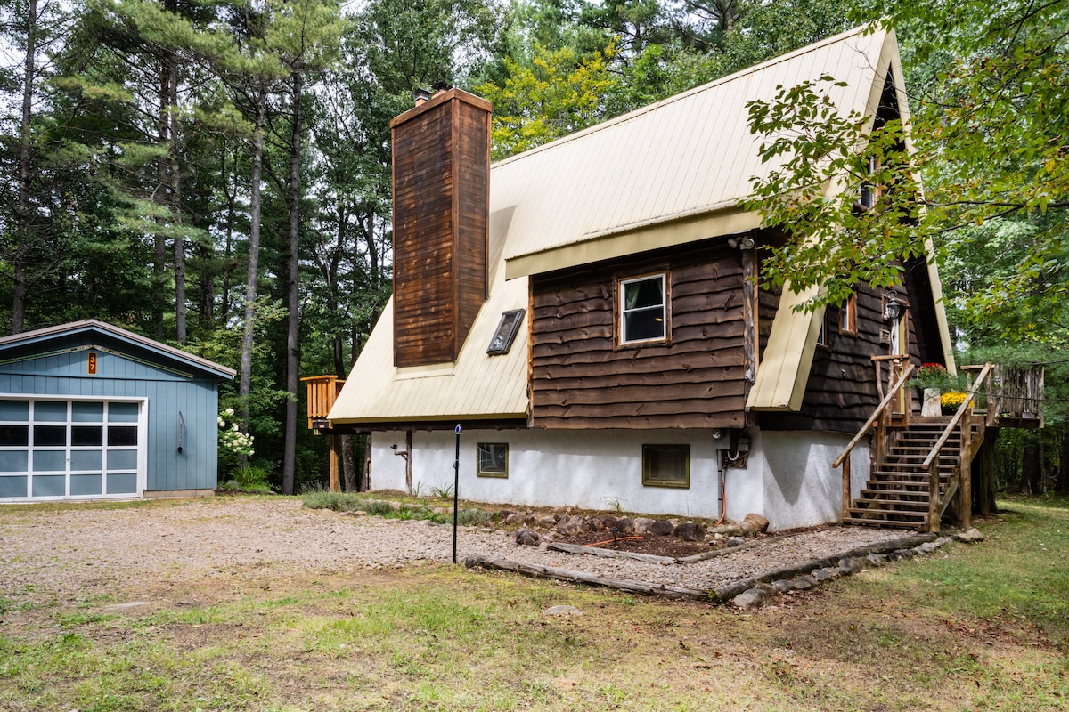 The exterior view of a cozy A-frame cabin is presented, featuring a pitched roof with a metal overhang and a stone chimney. The structure is set amidst trees, with a gravel area in front and a blue garage to the left. A wooden staircase leads to a side entrance.