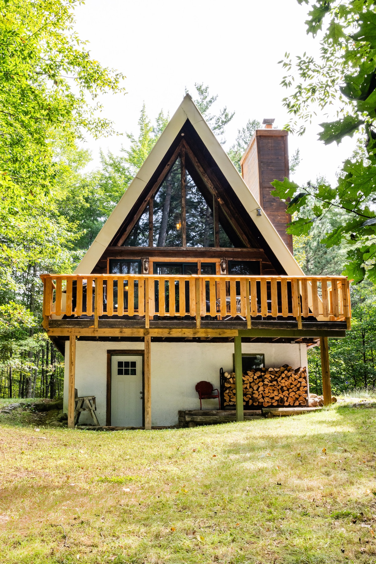 An A-frame cabin is captured amidst lush greenery, featuring a wall of windows that allow natural light to fill the interior. The spacious deck is accessible from the upper level, while wood logs are neatly stacked under the deck for outdoor use.