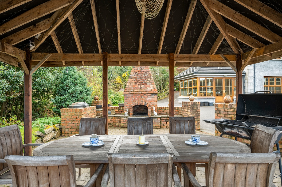 An outdoor dining area is featured under a wooden gazebo, with a large round table surrounded by six wooden chairs. A brick fireplace and a barbecue grill are visible in the background, complemented by lush greenery and a stone pathway.