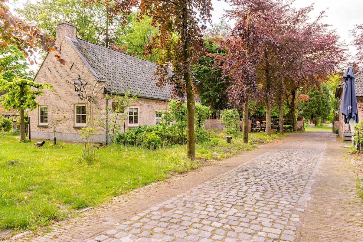 A charming building with light-colored stone walls is framed by lush greenery and flowering trees. A cobblestone pathway leads to the entrance, creating a serene atmosphere. The surrounding area features a well-maintained garden, enhancing the welcoming appeal of the property.