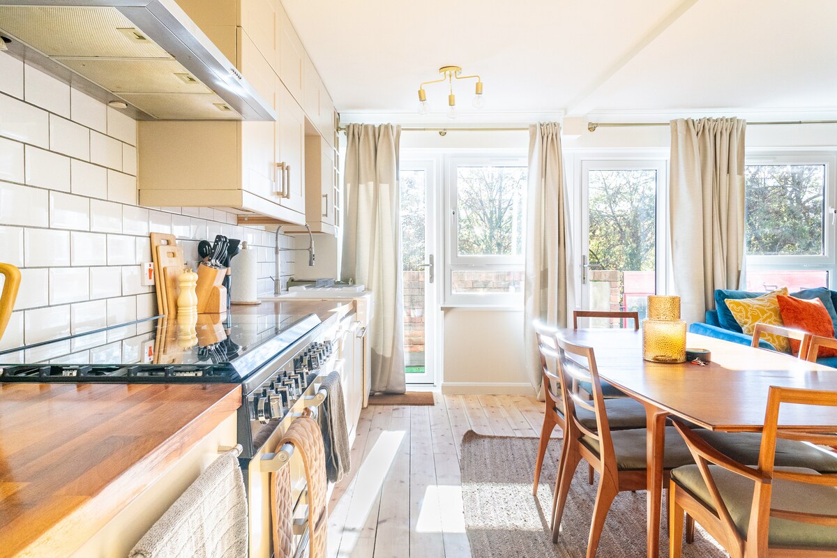 The open kitchen features sleek cabinetry and a gas range, complemented by a wooden dining table surrounded by chairs. Natural light fills the room through large windows, with warm tones from the furnishings and decor enhancing the inviting ambiance.