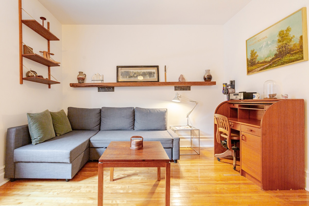 A spacious living area is featured, showcasing a gray sectional sofa accompanied by two green accent pillows. A wooden coffee table sits centrally, while a desk and lamp are positioned in the corner. Shelves display various decorative items, and a piece of artwork adorns the wall.