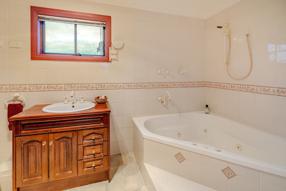 An ensuite bathroom features a spacious double spa with jets, complemented by a wooden vanity with several storage drawers. A window above provides natural light, while tiled walls and floor create a clean, neutral atmosphere. A showerhead is mounted on the wall above the spa.