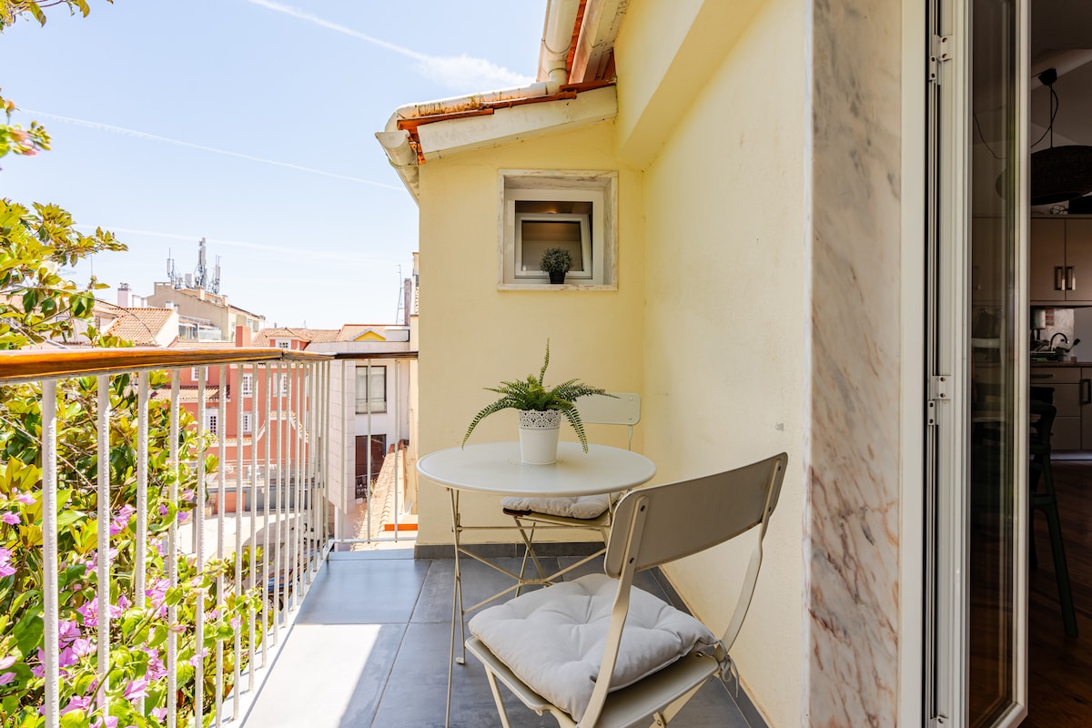 A small outdoor balcony features a round table with a potted plant and two foldable chairs. Light yellow walls and a view of nearby buildings are visible, creating a bright and airy space for relaxation.