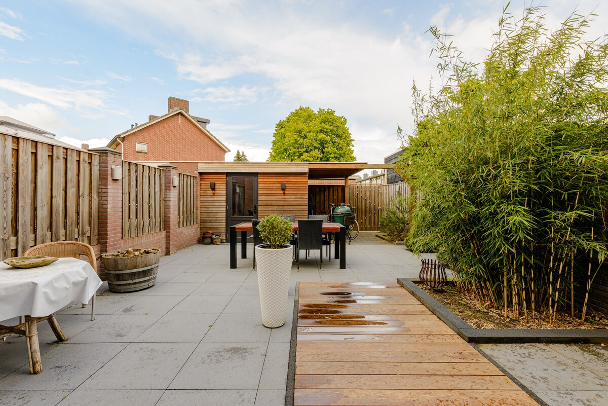 An inviting outdoor space is featured, showcasing a wooden deck leading to a patio area with a dining table and chairs. A barbecue is positioned nearby, surrounded by greenery and bamboo for a touch of nature. Paved stones add a modern feel to the area.