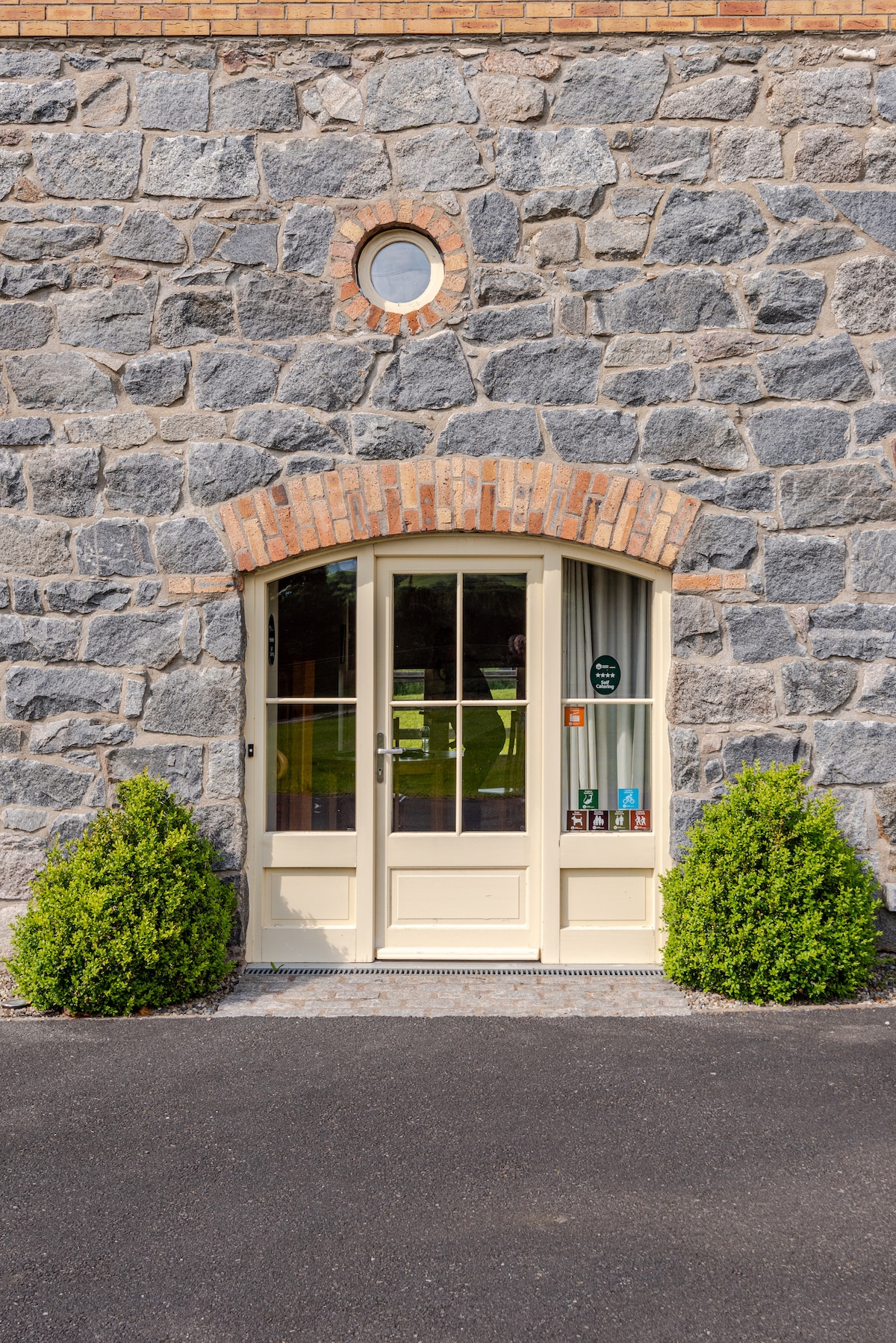 The barn's entrance is framed by a natural stone wall, showcasing a double door with clear glass panels. Flanking the door, two neatly trimmed green shrubs provide a touch of greenery. Above, a circular window adds an architectural element to the facade.