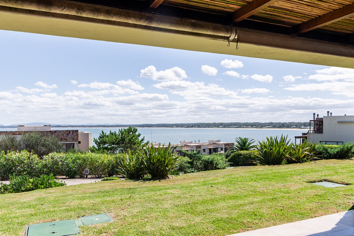 A spacious outdoor area is showcased, framed by a covered terrace. An expansive view of the calm waters is visible, complemented by lush green landscaping and neighboring buildings. The clear sky contributes to a peaceful atmosphere.