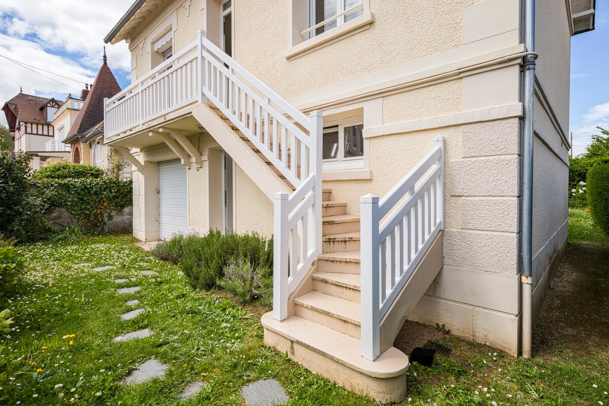 A set of white stairs leads to the entrance of the house, flanked by a protective railing. The pathway is bordered by green grass and a few blooming flowers, contributing to the home's welcoming exterior.