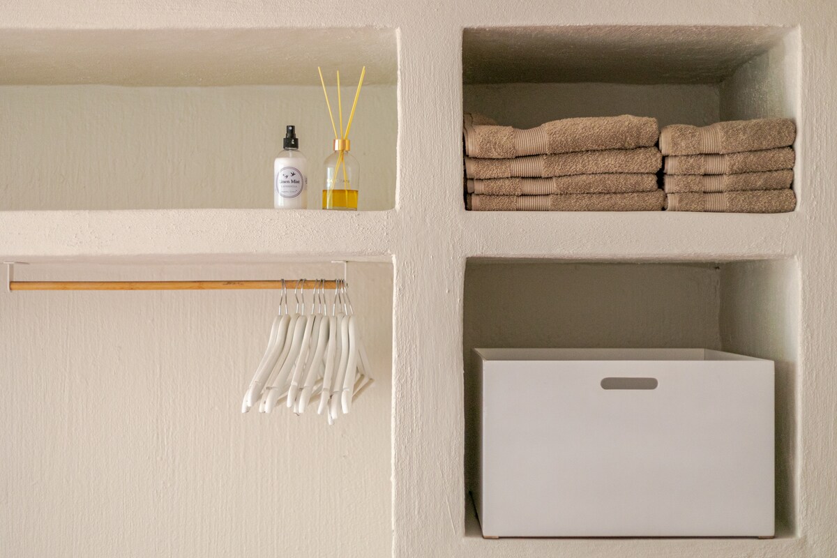 A neat shelving unit displays rolled towels and bath products, including a bottle of lotion and a diffuser. Wooden hangers are arranged on a minimalistic rod, with a storage box situated below for additional organization.