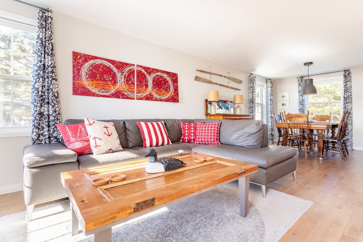 A spacious living area features a large sectional sofa adorned with patterned cushions and a rustic coffee table. Two pieces of colorful artwork hang on the wall, complemented by decorative items on the table. The dining table can be seen in the background, surrounded by wooden chairs.