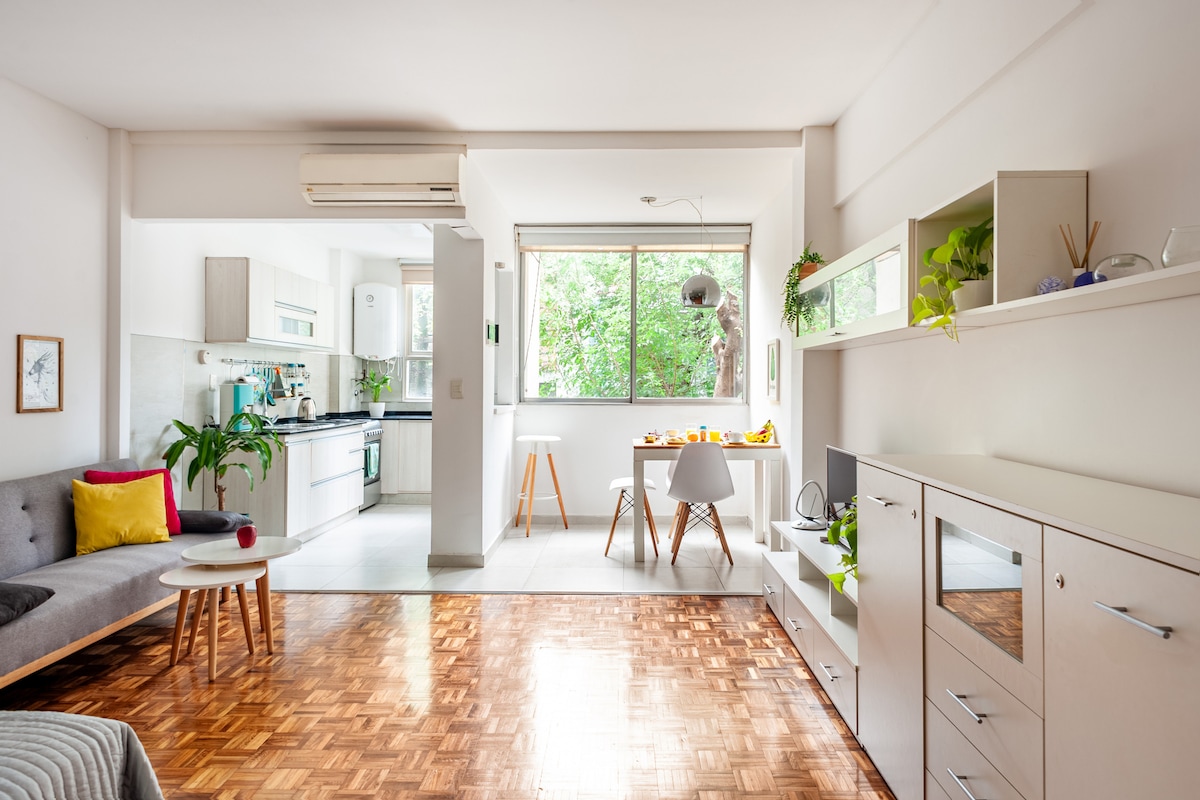 A bright living area is presented with wooden flooring, featuring a cozy sofa adorned with colorful cushions. A dining table with modern chairs is arranged beside a large window, allowing natural light to illuminate the space. The kitchen is visible, showcasing clean lines and a functional layout.