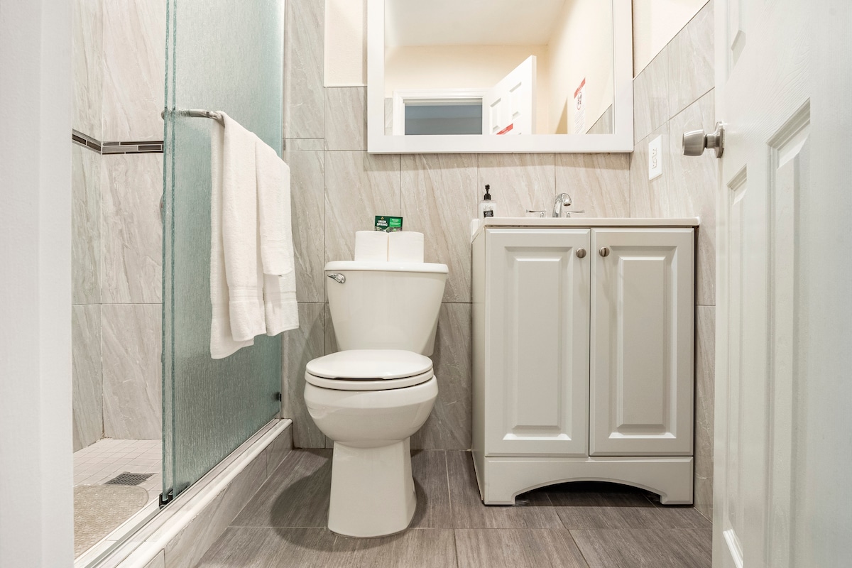 A modern bathroom is displayed, featuring a glass-enclosed shower, a white toilet, and a vanity with storage cabinets. Light gray tiles are used for the flooring and walls. A mirror is mounted above the sink, and a towel hangs neatly on a bar beside the shower.