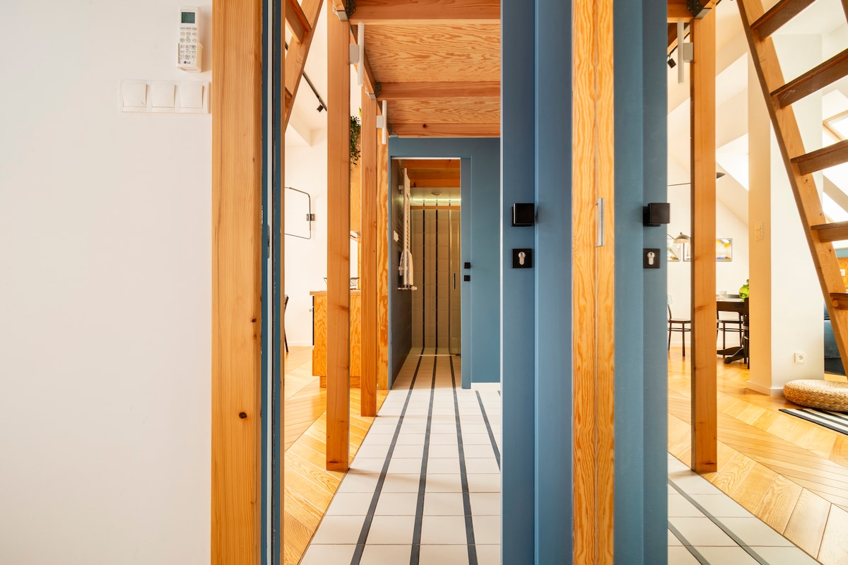 A spacious hallway is highlighted by warm wood beams and chevron wood flooring. The walls are painted in soothing tones, providing a serene backdrop. Two mirrored doors lead to rooms, while soft lighting enhances the welcoming environment. A staircase is visible in the background.