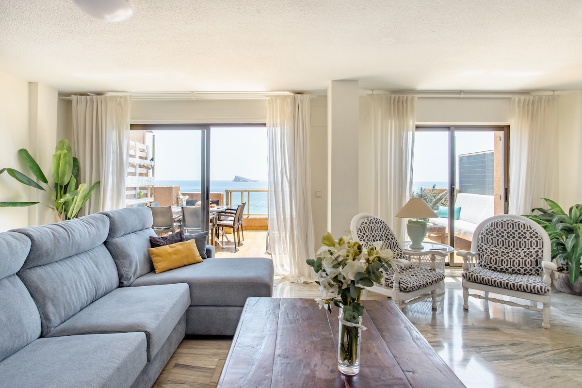 A bright living area is furnished with a spacious grey sofa and a wooden coffee table. Large glass doors open to a balcony overlooking the sea, while white curtains gently diffuse the light. Potted plants and decorative chairs enhance the inviting atmosphere.