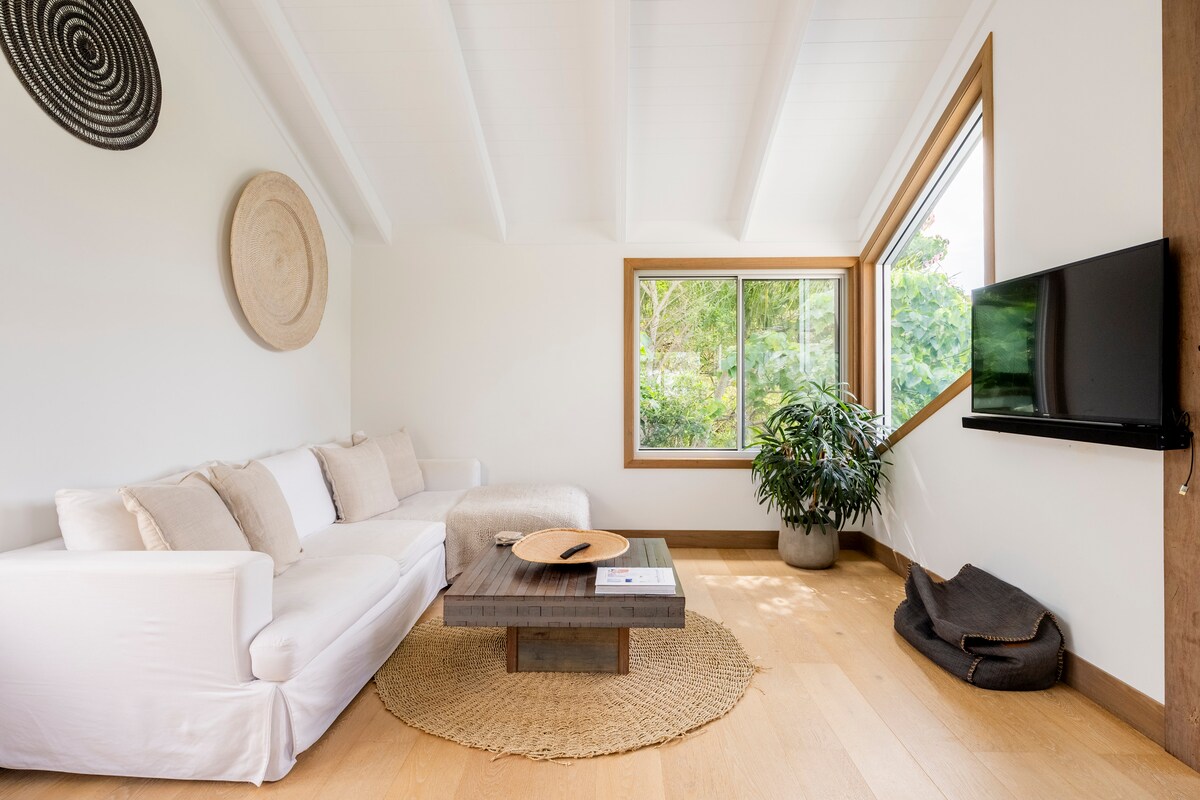 A bright, airy lounge area features a cozy white sectional sofa positioned next to a natural wood coffee table. A large window fills the space with light, offering views of greenery. Decorative items include a woven wall hanging and a potted plant, enhancing the welcoming atmosphere.