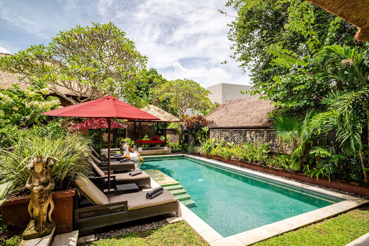 A serene outdoor space features a 10m x 4m private swimming pool surrounded by lush tropical gardens. Sun loungers with umbrellas provide comfortable seating, while traditional Balinese statues add local charm. The covered gazebo offers a shaded area for relaxation.