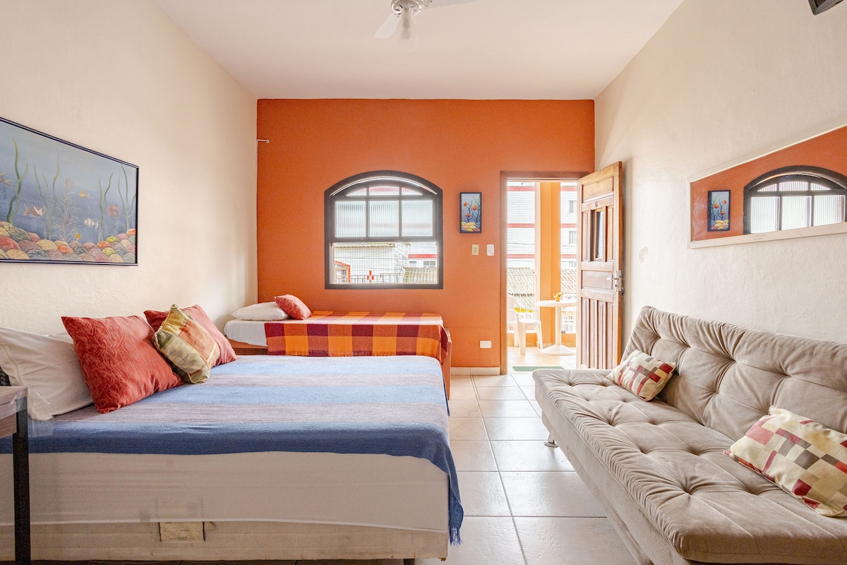 A bright room features a spacious double bed, a single bed, and a comfortable sofa bed. Soft pillows in various colors are arranged on the beds. A colorful artwork adorns one wall, and a large window offers views into a sunny outdoor space.