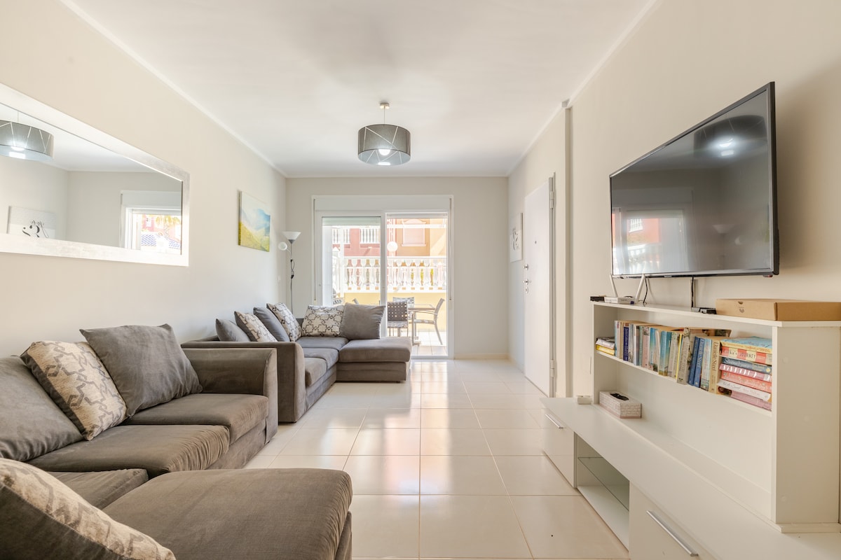 A bright living area features comfortable sofas arranged for relaxation. A large television is mounted on the wall, and a mirrored accent reflects the light-filled space. Patio doors open to an outdoor seating area, enhancing the connection between indoor and outdoor living.