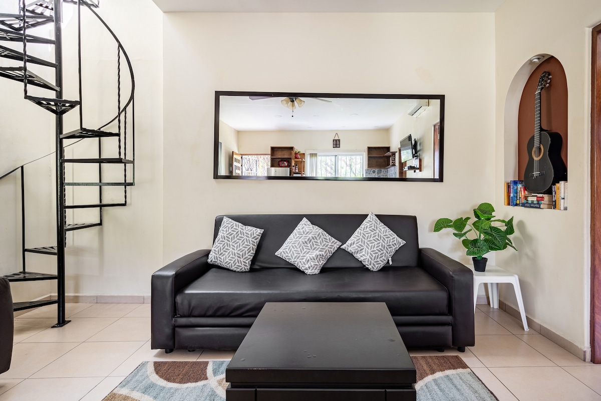 A modern living area features a dark sofa adorned with decorative pillows, complemented by a coffee table. A spiral staircase leads to the upper level, and a large mirror reflects the space. A guitar is displayed on the wall, adding to the relaxed ambiance.