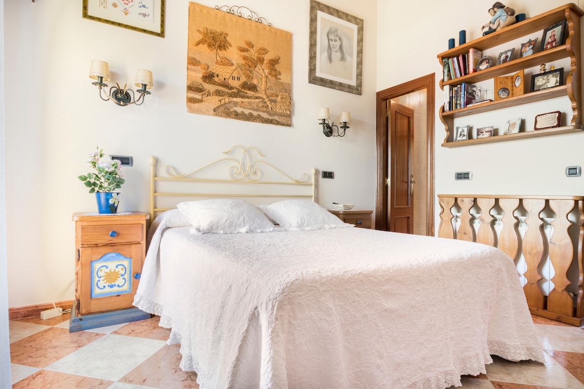 A cozy bedroom is presented with a light-colored bedspread covering a spacious bed. Decorative wall art and a wooden shelf filled with books and framed photos add character. Soft lighting from sconces enhances the inviting atmosphere, while a small dresser beside the bed features floral decor.