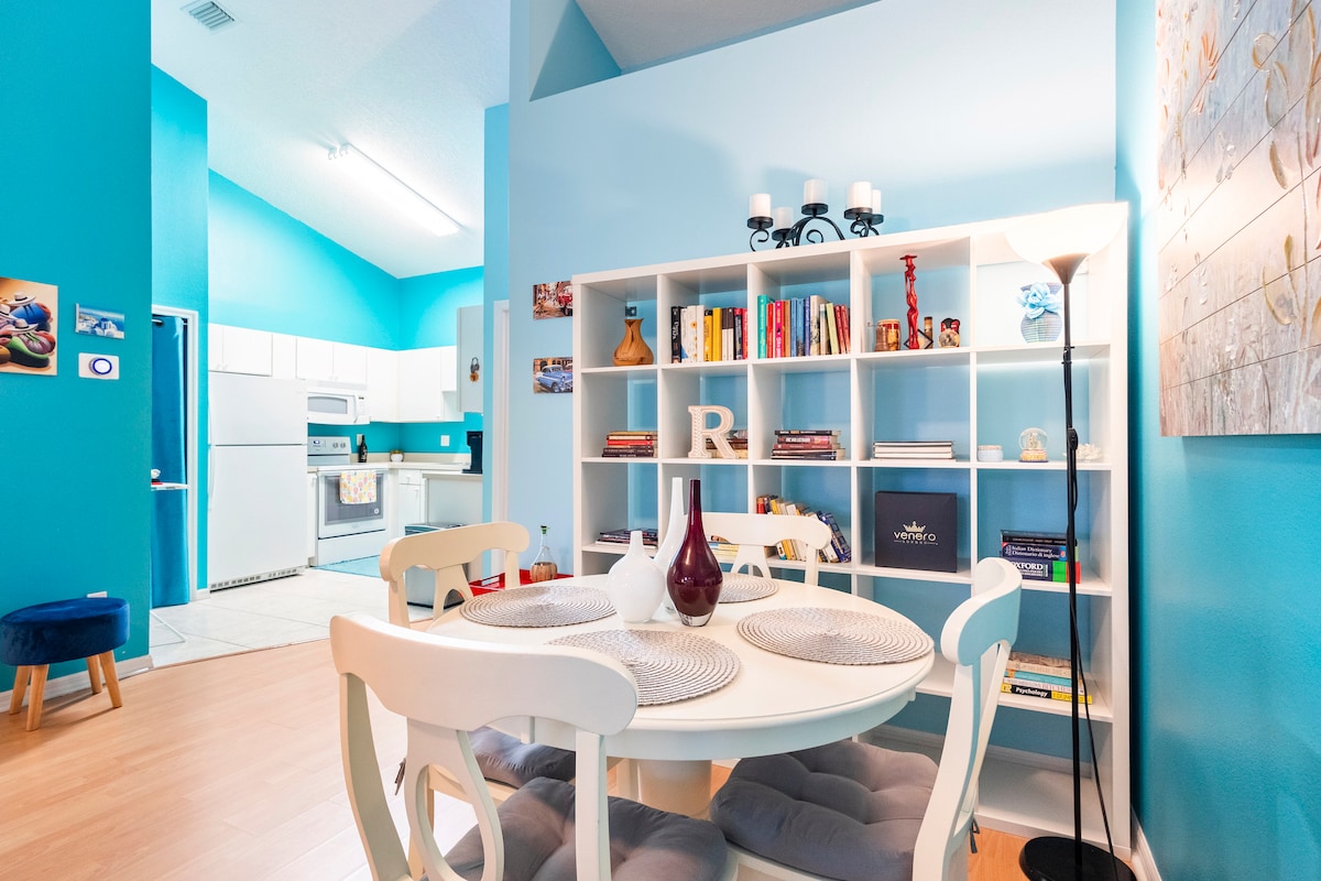 A light-filled dining area features a round table surrounded by four white chairs, complemented by a decorative bookshelf showcasing various books and decor. A floor lamp stands nearby, illuminating the space, while a glimpse of the modern kitchen can be seen in the background.
