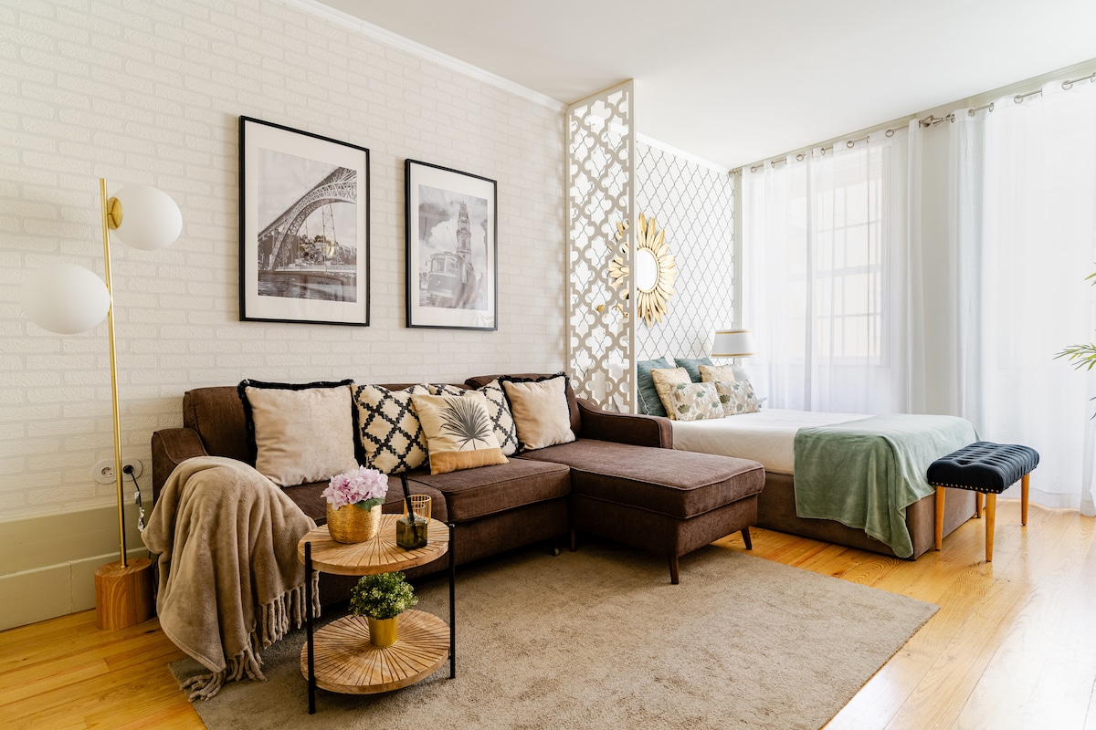 A spacious living area is furnished with a comfortable sofa and decorative cushions. A double bed is positioned nearby, framed by soft curtains. Two framed photographs are displayed on the light-colored wall, while a small coffee table with plants enhances the inviting ambiance.