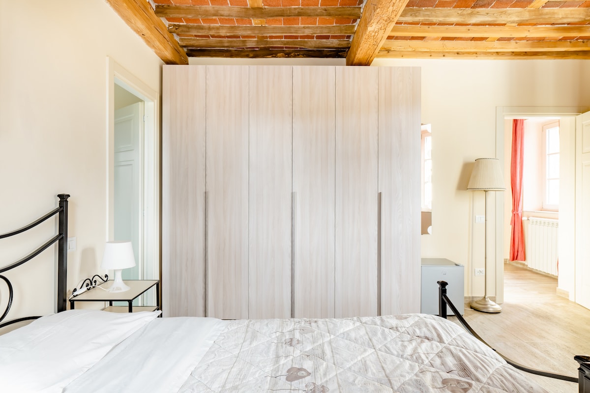 A spacious room is shown, featuring a large bed with a light-colored quilt and a black metal frame. A modern wardrobe with vertical panels is positioned against the textured ceiling. A bedside table with a lamp sits beside the bed, and soft natural light enters through a nearby doorway.