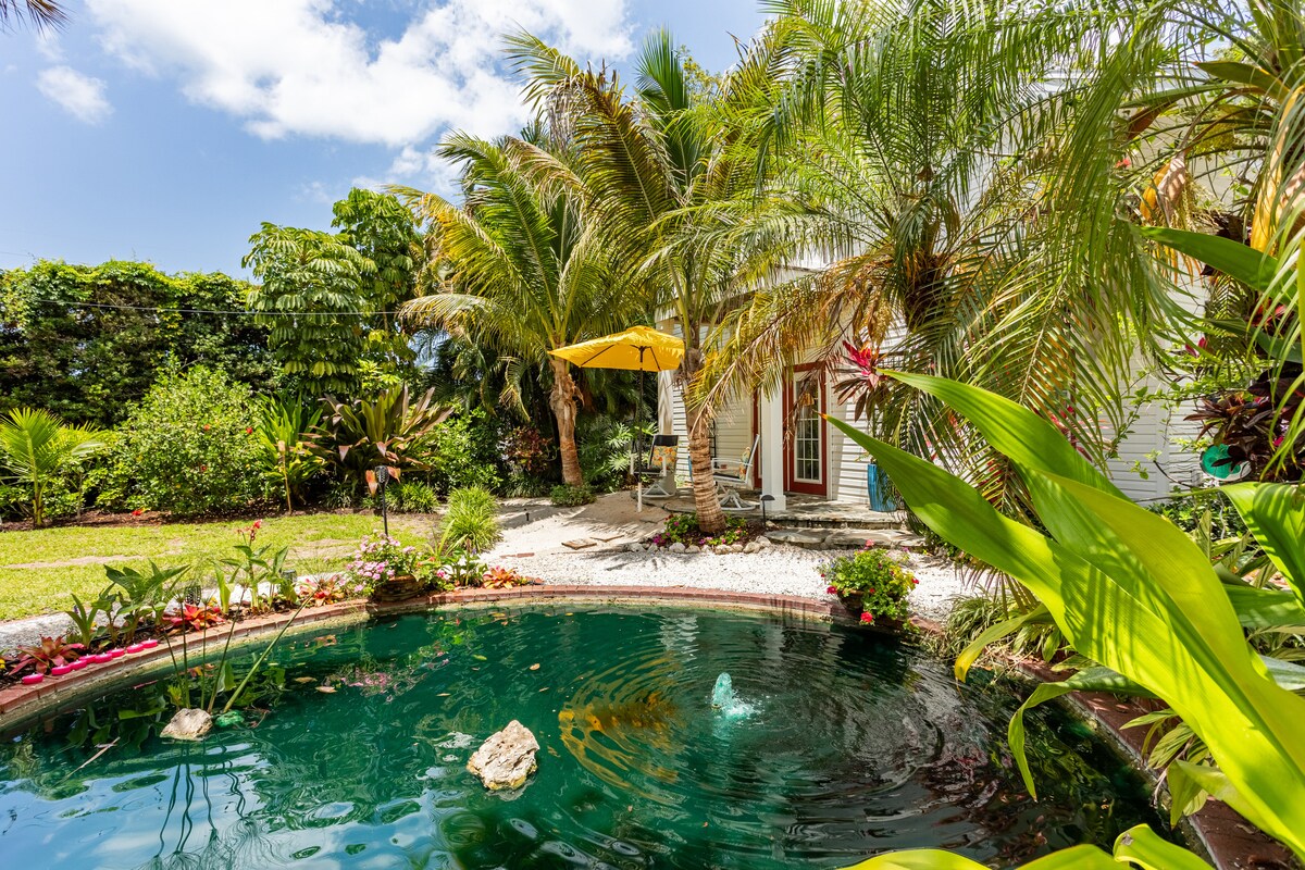 A tranquil oasis is set within vibrant greenery, with a refreshing lagoon pool surrounded by tropical plants. An umbrella provides shade over the outdoor seating area, while colorful flowers add detail to the landscaped surroundings, inviting relaxation and a connection with nature.